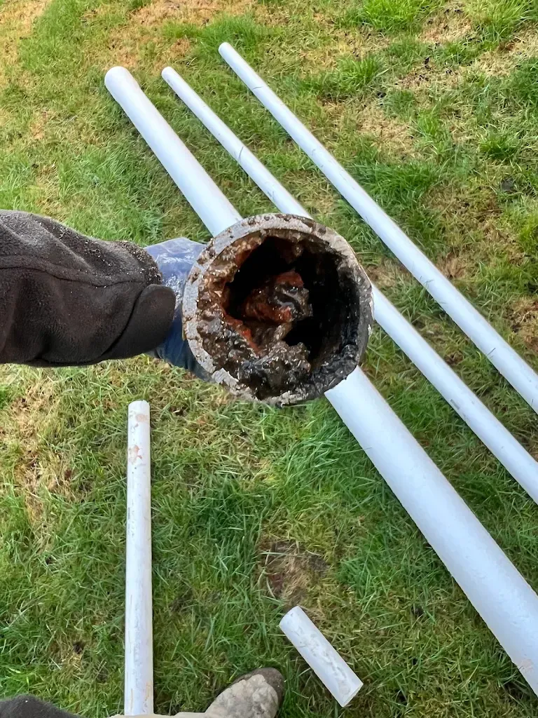 Hand holding a cut section of old drain pipe nearly blocked with debris, with new PVC pipe laid out on the lawn