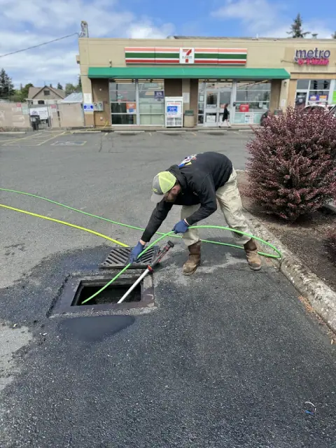 Plumber hydro jetting a parking lot storm drain catch basin outside a convenience store
